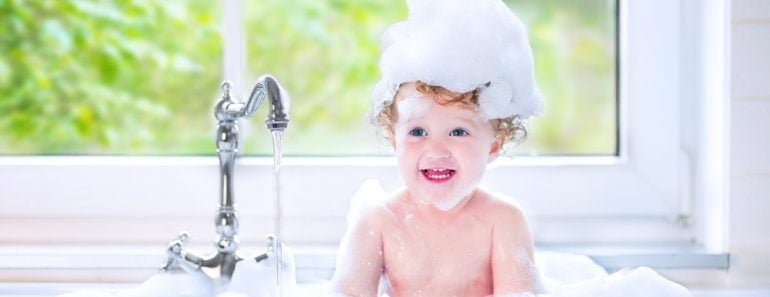 Funny little baby girl with wet curly hair taking a bath in a kitchen sink with lots of foam playing with water drops and splashes next to a big window with garden view