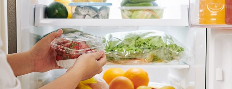 Hands of woman opening fridge door and putting package of fresh ripe strawberries in it